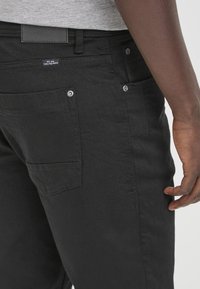 Črne jeans hlače z gladko teksturo, petimi žepi in srebrnimi dodatki. Imajo oznako na hrbtni strani, kar nakazuje na vsakodnevno nošenje.