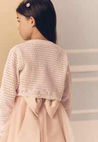Cardigan rosa lavorato a maglia con un motivo all'uncinetto e orlo a ondine, abbinato a un vestito leggero rosa chiaro con un grande fiocco di satin sulla schiena.