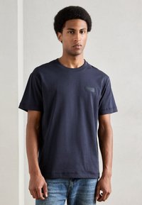 Σκούρο μπλε κοντομάνικο μπλουζάκι από βαμβάκι με στρογγυλή λαιμόκοψη και μικρή μαύρη λογότυπη επιφάνεια στο αριστερό στήθος. Φοριέται με ανοιχτόχρωμα τζιν.