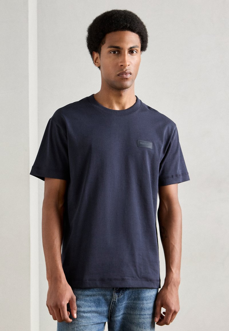 Σκούρο μπλε κοντομάνικο μπλουζάκι από βαμβάκι με στρογγυλή λαιμόκοψη και μικρή μαύρη λογότυπη επιφάνεια στο αριστερό στήθος. Φοριέται με ανοιχτόχρωμα τζιν.