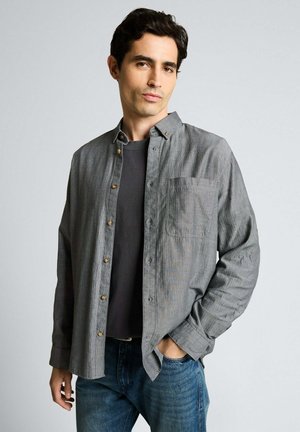 Jeune homme aux cheveux foncés portant une chemise à rayures grise boutonnée sur un t-shirt noir et un jean bleu, une main dans la poche, fond neutre.