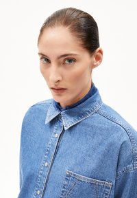 Blue denim shirt with a classic collar, front pocket, and visible stitching details. Smooth texture and button closures at the collar.