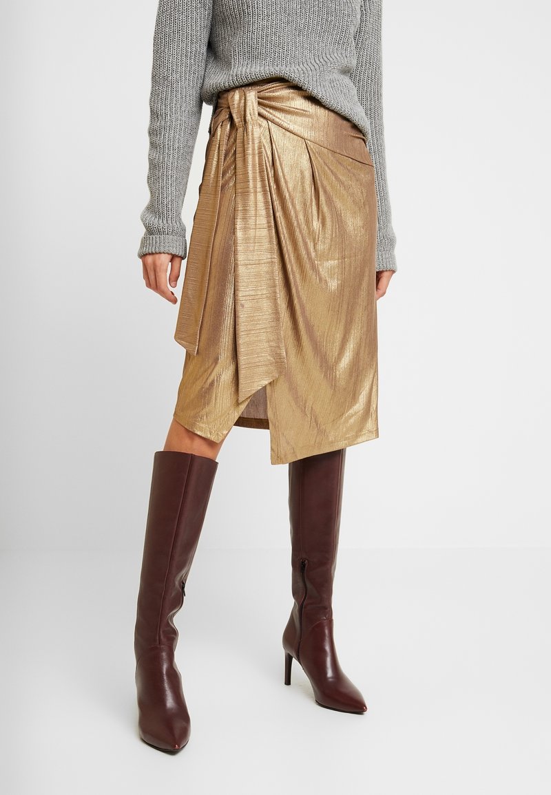 Guldig metallisk omslagskjol med plisserad design och draperade detaljer, kombinerad med knähöga burgundyläder stilettklackar. Grå stickad tröja.