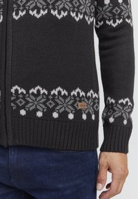 Dunkelgrauer Strickcardigan mit weißen und grauen Schneeflockenmuster. Verfügt über gerippte Bündchen und Saum sowie ein kleines Markenetikett.