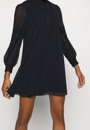 Vestido mini negro de manga larga con mangas transparentes, detalles de encaje en la parte delantera y los puños, llevado por una persona sobre un fondo liso.