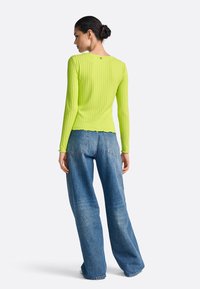 Testhez simuló, bordázott hosszú ujjú felső neon zöld színben, fodros szegéllyel és kerekített hátrésszel. Laza, világoskék farmernadrággal párosítva.