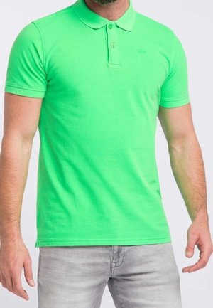 Schmal geschnittenes, knallgrünes Poloshirt mit kurzen Ärmeln und Zwei-Knopf-Leiste, getragen mit hellgrauen Jeans an einem stehenden männlichen Modell.