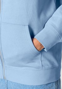 Felpa azzurro chiaro con zip frontale, polsini a costine e tasca a marsupio. Tessuto dalla trama liscia con vestibilità comoda.