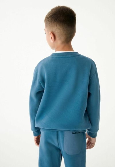 Felpa blu e pantaloni coordinati in morbido tessuto, con polsini a costine e una tasca sul retro con dettaglio del logo. Taglio semplice e comodo.