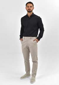 Zwarte button-up shirt, beige broek en lichtgrijze instappers. Het shirt heeft een effen textuur en de broek heeft een klassieke pasvorm.