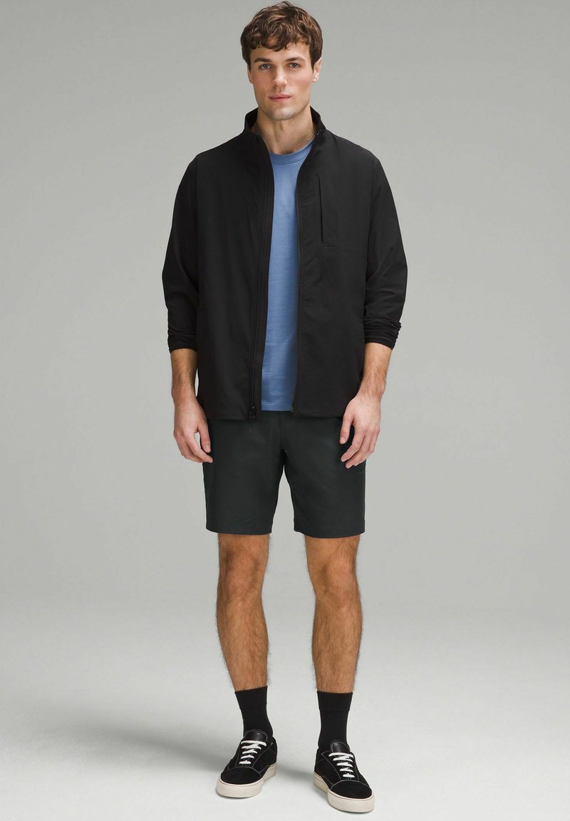 Czarna kurtka zapinana na zamek na niebieskiej koszuli, w połączeniu z ciemnymi szortami i czarnymi trampkami. Wyeksponowana na neutralnym tle.