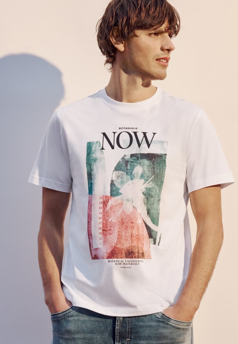 Homme portant un T-shirt blanc graphique avec un dessin abstrait de plante et les mots « NOW » et « Botanical engineering raw materials » regardant sur le côté.