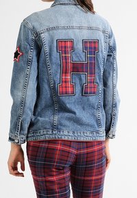 Denimjacka med en stor rutig "H"-patch på baksidan, samt en stjärnpatch på vänster ärm. Blå färg, standardkrage och knäppning.