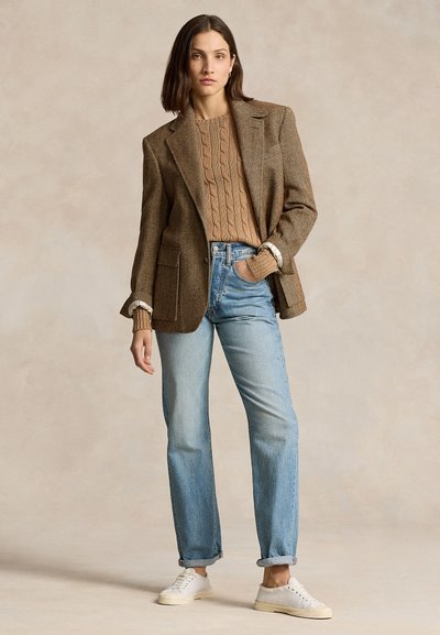 Sieviete stāv ar roku kabatā, valkājot brūnu adītu džemperi ar pinumu rakstu, brūnu zivisvara raksta žaketi, gaiši zilas džinsas un baltas sporta kurpes.