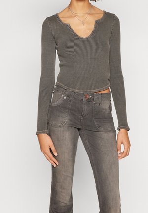 Frau trägt ein eng anliegendes, geripptes Langarmshirt mit wellenförmigem Ausschnitt und hoch taillierte, ausgebleichte graue Jeans, steht mit entspannten Armen.