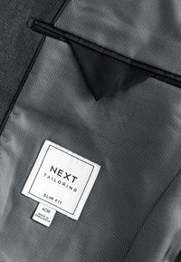 Tissu en mesh gris avec une étiquette de vêtement blanche portant la mention "NEXT TAILORING SLIM FIT 40R MADE IN BANGLADESH" à l'intérieur d'un vêtement.