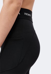 Close-up of a person wearing black high-waist Techfit leggings with visible stitching and a side pocket.