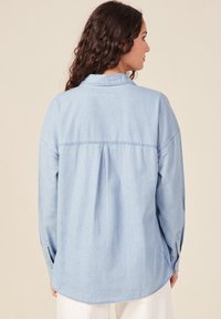 Camisa de mezclilla azul claro, de corte holgado, con mangas largas y puños abotonados, detalle de plisado en la parte posterior y un cuello clásico. Textura suave y diseño casual.
