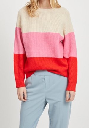 Person wearing a color-blocked sweater with cream, pink, and red stripes, paired with light blue high-waisted trousers, standing.