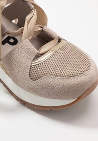 Sneaker beige con tomaia in mesh e dettagli in suede testurizzato, caratterizzata da un bordo dorato e una suola bianca spessa con suola esterna marrone.