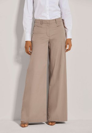 Beige wijde broek met een gladde textuur, hoge taille en riemlussen, gecombineerd met een witte blouse met lange mouwen en neutrale sandalen.