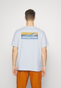 Gaiši zils kokvilnas T-krekls ar taisnstūra grafiku uz aizmugures, kurā attēlotas viļņu formas un vārds "patagonia" baltā krāsā. Īsas piedurknes, brīva piegriezuma.