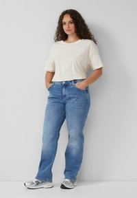 T-shirt beige clair, coupe ample, associé à un jean bleu taille moyenne, jambe droite, délavé, porté avec des baskets de sport argentées.