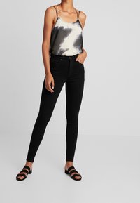 Jeans ajustados negros combinados con una blusa sin mangas de tirantes en color gris y crema con un diseño de tie-dye, que presenta tirantes finos y un corte relajado. Sandalias negras con tiras.