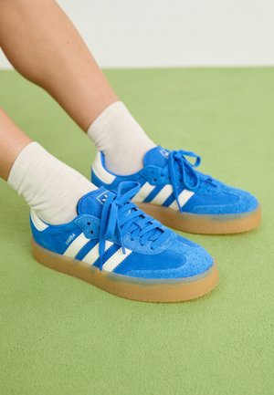 Pieds portant des baskets Adidas Samba bleu vif avec des rayures blanches et des semelles beiges sur un tapis vert clair.