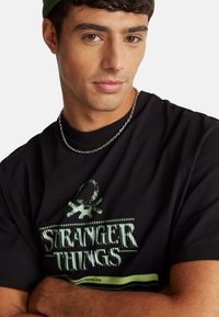 Černé tričko se zeleným potiskem "Stranger Things", kulatým výstřihem, krátkými rukávy a stříbrným řetízkovým náhrdelníkem. Texturovaný bavlněný materiál.