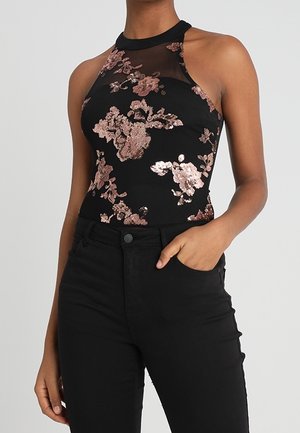 Czarna bluzka bez rękawów z prześwitującym dekoltem i różowo-złotymi kwiatowymi cekinami, noszona z wysokopasmowymi czarnymi spodniami, jedna ręka w kieszeni.