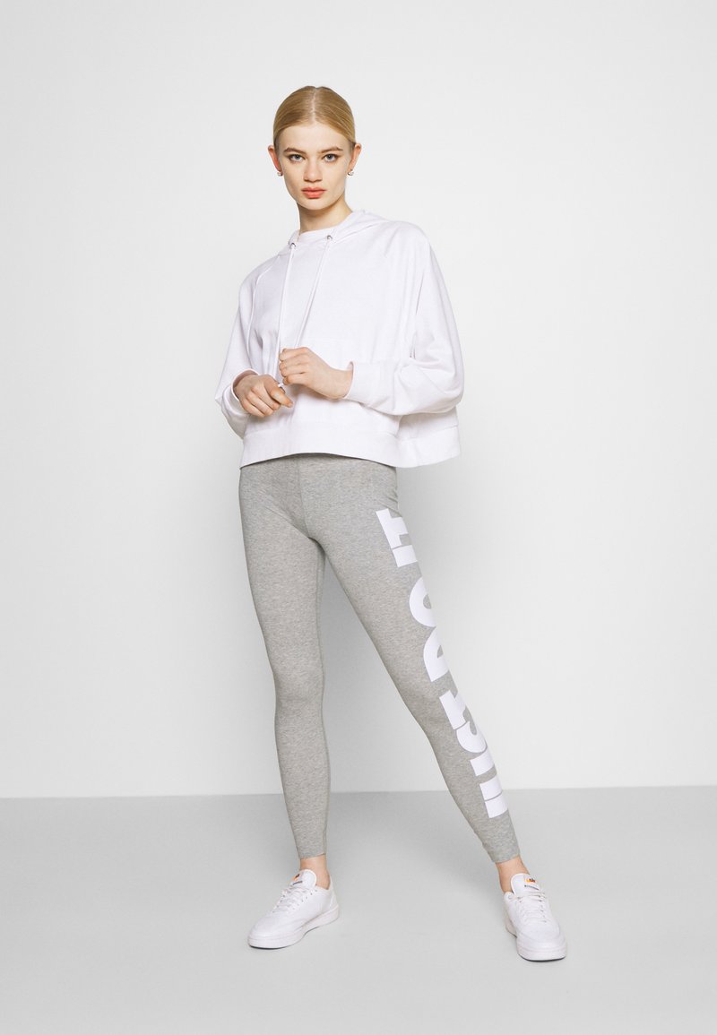 Sudadera blanca corta con cordones, combinada con leggings grises que tienen un gran texto blanco en el costado. Zapatillas blancas completan el atuendo.