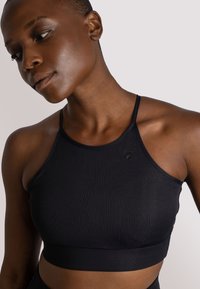 Crop top sportivo nero a coste con spalline sottili, caratterizzato da un design aderente e un accento logo discreto. Texture liscia, ideale per l'abbigliamento sportivo.