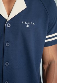 Marineblauwe polo met een contrasterende lichtgekleurde kraag, een twee-knoopsluiting en witte streepaccenten op de mouwen. Merkenlogo geborduurd.