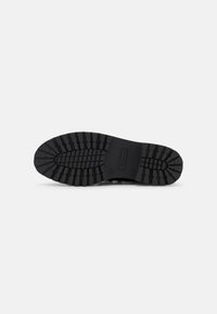 Semelle de chaussure en caoutchouc noir avec un motif de texture, comprenant des rainures et des crêtes pour une meilleure adhérence, et une zone centrale pour le logo de la marque.