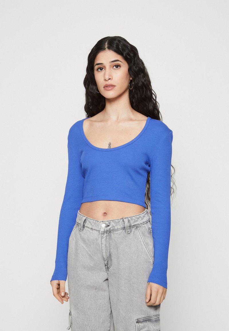 Dugi rukavi plavi crop top s rebrastom teksturom, uparen s visokim sivim trapericama. Ima široki izrez i prilagođeni dizajn.