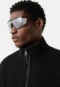 Sort fleecejakke med høj krave, lynlås med sølvdetalje, kombineret med reflekterende solbriller.