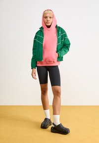 Zelená zip-up bunda s bielymi akcentmi, ružová mikina s kapucňou, čierne biker šortky a čierne crocs. Modelka stojí na žltej podlahe pred svetlým pozadím.