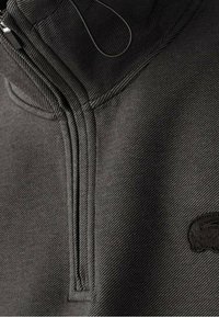 Veste noire légère avec fermeture éclair et col montant. Dispose d'un tissu texturé, de coutures subtiles et d'un petit logo sur la poitrine.