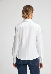 White long-sleeve shirt with a collar, featuring a straight back seam and cuff buttons, paired with dark blue jeans. Smooth fabric texture.