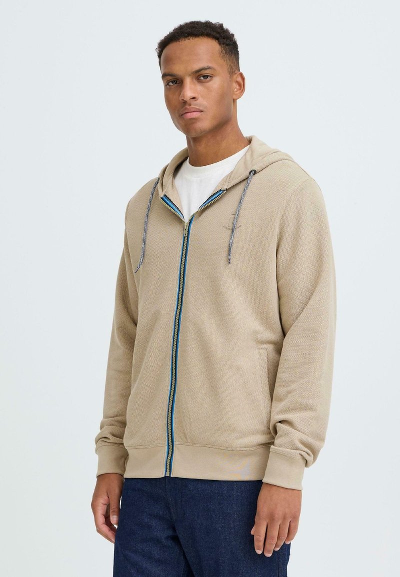 Sweat à capuche beige à fermeture éclair avec un tissu texturé, une fermeture éclair bleue contrastante et des cordons de capuche. Dispose de poches latérales et de poignets côtelés.