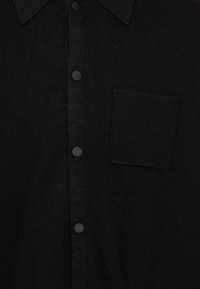 Chemise noire à boutons avec une petite poche poitrine et un col pointu, comportant des boutons ronds noirs mats sur le devant.