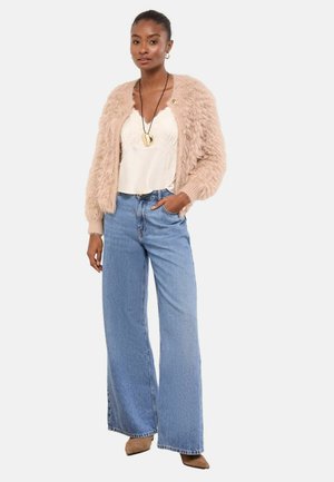 Fuzzy beige cardigan over a white lace cami, paired with wide-leg blue jeans. Notable gold pendant necklace and light brown shoes.