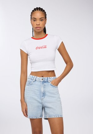 T-shirt bianca cropped con colletto rosso e logo Coca-Cola, abbinata a pantaloni shorts di jeans azzurri a vita alta.