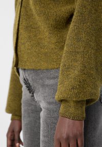 Ελιά πράσινο πλεκτό cardigan με απαλό υφασμά, ριχτές μανσέτες και ίσια στρίμωξη, συνδυασμένο με γκρι υφασματισμένα τζιν.