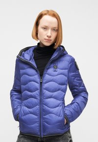 Femme aux cheveux rouges coupés au carré, portant une veste matelassée bleue à capuche par-dessus un col roulé noir, les mains dans les poches.