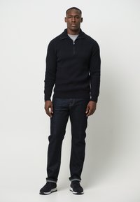 Granatowy sweter dzianinowy z kołnierzem na pół zamek, zestawiony z ciemnoniebieskimi dżinsami i czarnymi butami sportowymi. Prosty, teksturowany materiał z prążkowanym wzorem.