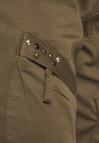 Tkanina olivově zelené barvy s boční kapsou, zdobenou mosaznými a černými kovovými hroty, prošívanými detaily a strukturovaným povrchem.