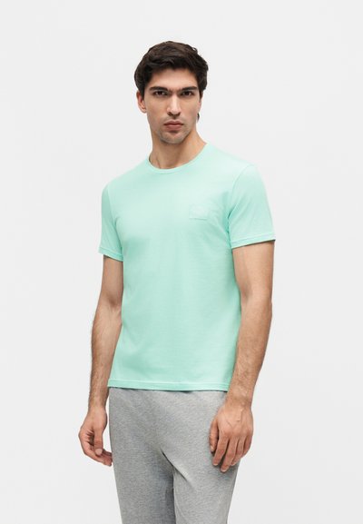 Νέος άντρας στέκεται φορώντας πράσινο μπλουζάκι κοντομάνικο και ανοιχτό γκρι φόρμα μπροστά σε απλό ανοιχτό φόντο.