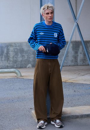 Jeune personne aux cheveux blonds courts portant un pull rayé bleu et noir, un pantalon large marron, et tenant une casquette bleu marine en plein air.